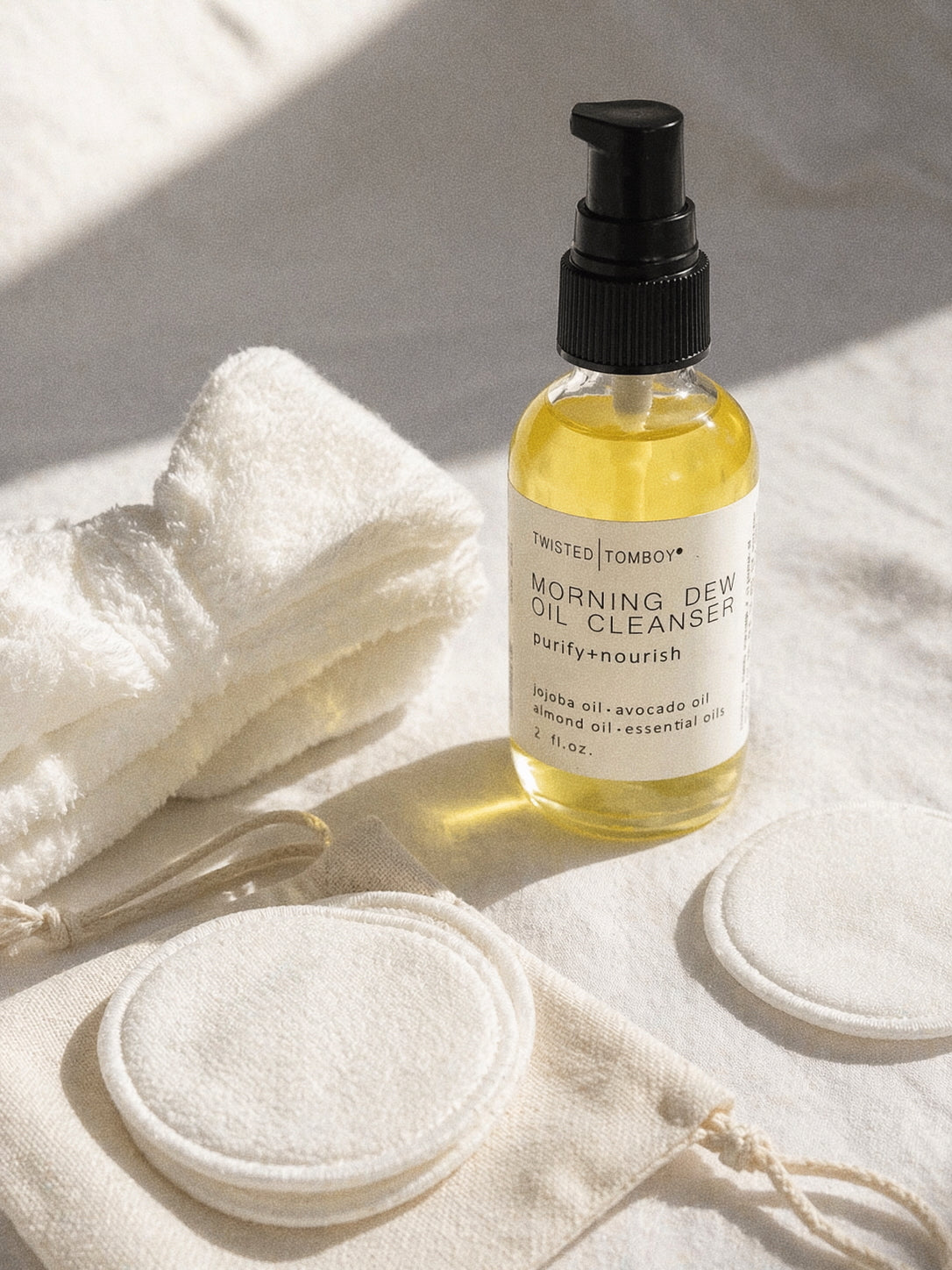 Bottle of 'Twisted Tomboy' morning dew oil cleanser on a white cloth with a cotton pad and towel.