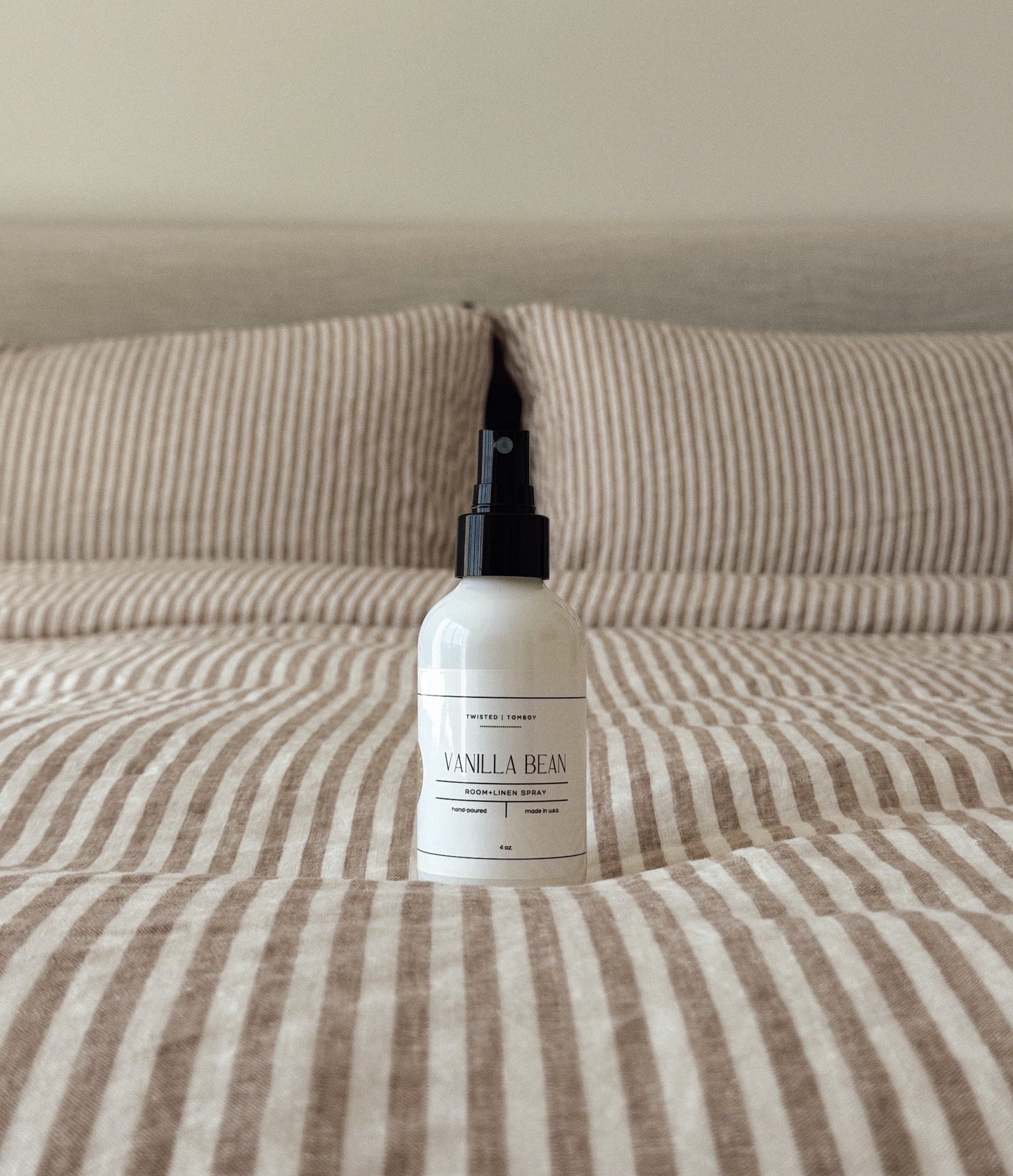 Vanilla bean room and linen spray bottle with black nozzle sitting on neutral striped bed linens